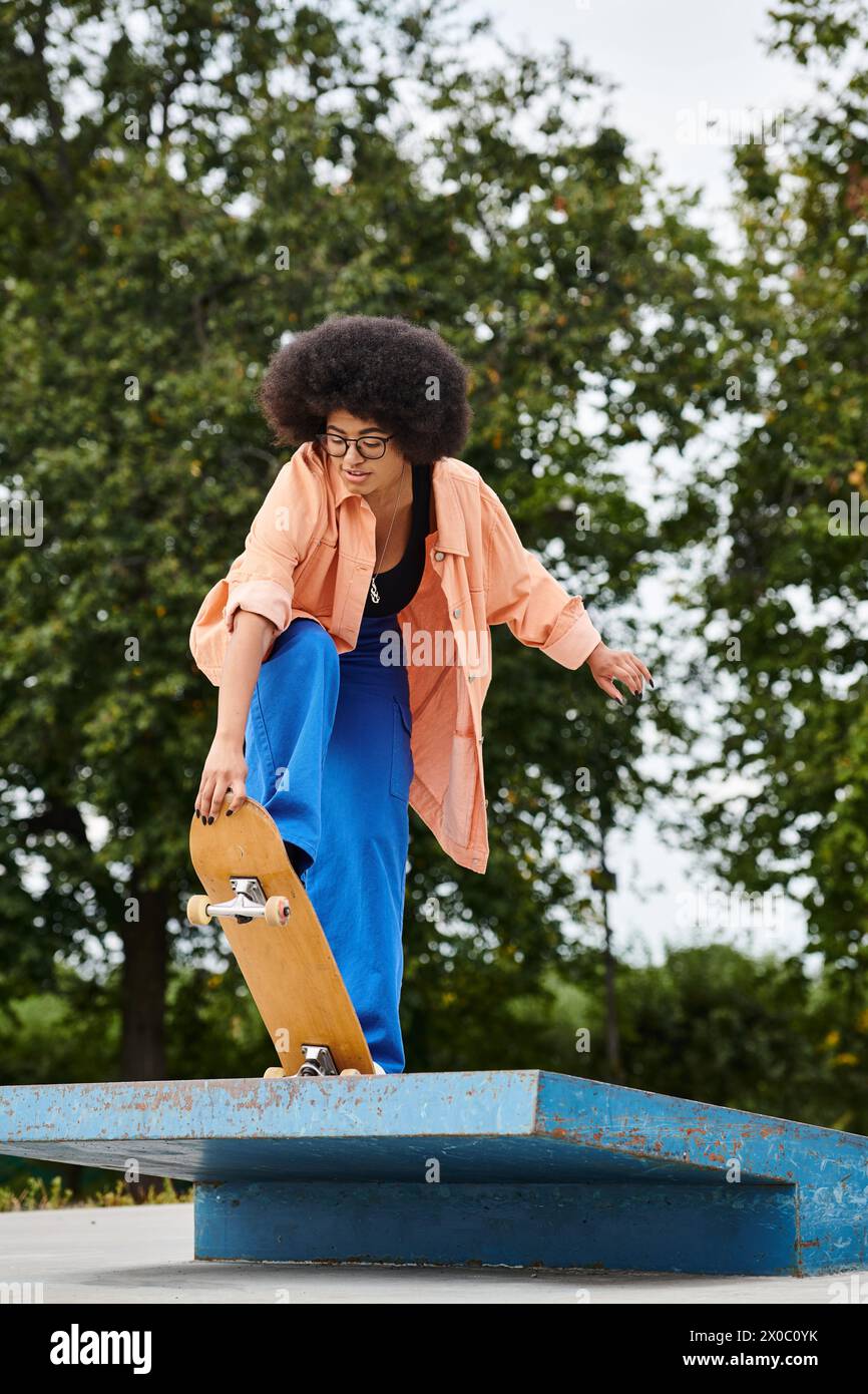 A young African American woman with curly hair, wearing blue pants and an orange shirt, executes a trick on a skateboard at a vibrant skate park. Stock Photo