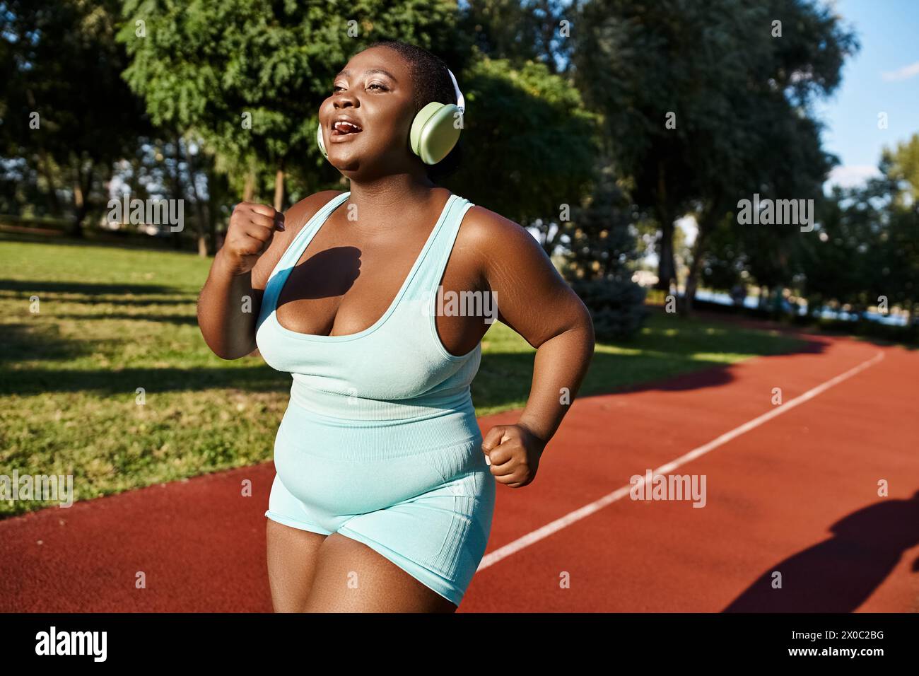 An African American woman in sportswear tunes into music with headphones while running on a track, embodying body positivity. Stock Photo
