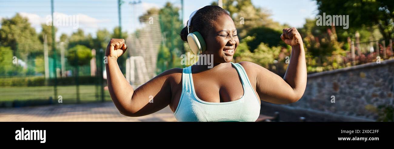 An African American woman in a sports bra top flexes her muscles confidently outdoors, showcasing body positivity and strength. Stock Photo