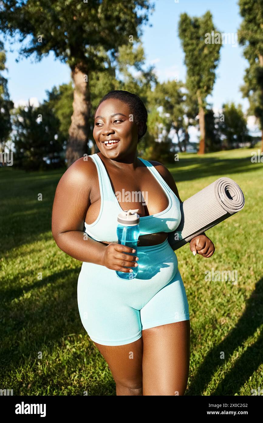 A joyful African American woman in sportswear, curvy and body positive, holds a yoga mat and a bottle of water outdoors. Stock Photo