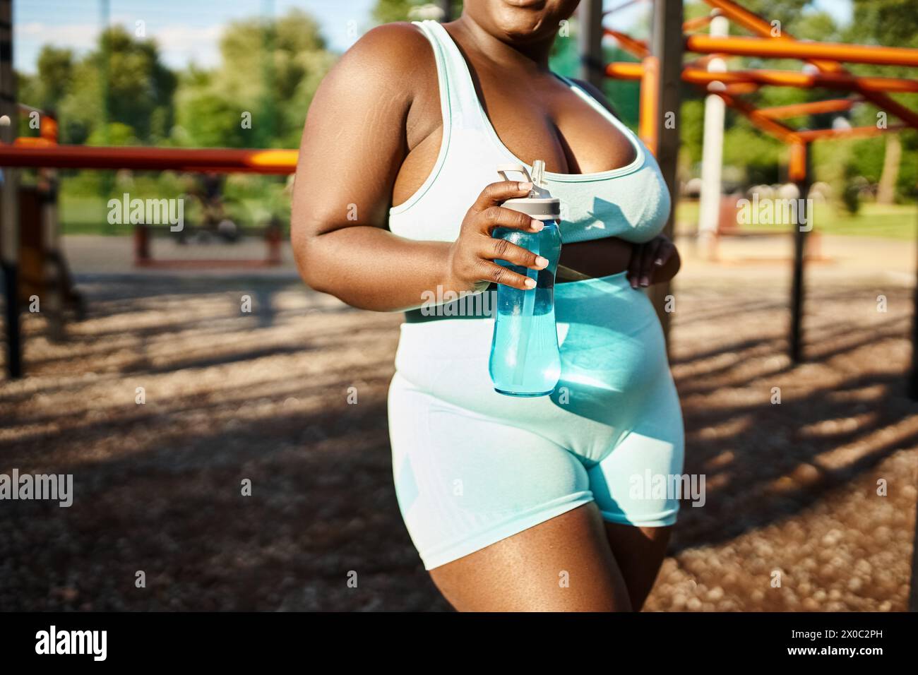 An African American woman in a white bodysuit, showcasing body positivity, holding a water bottle while exercising outdoors. Stock Photo