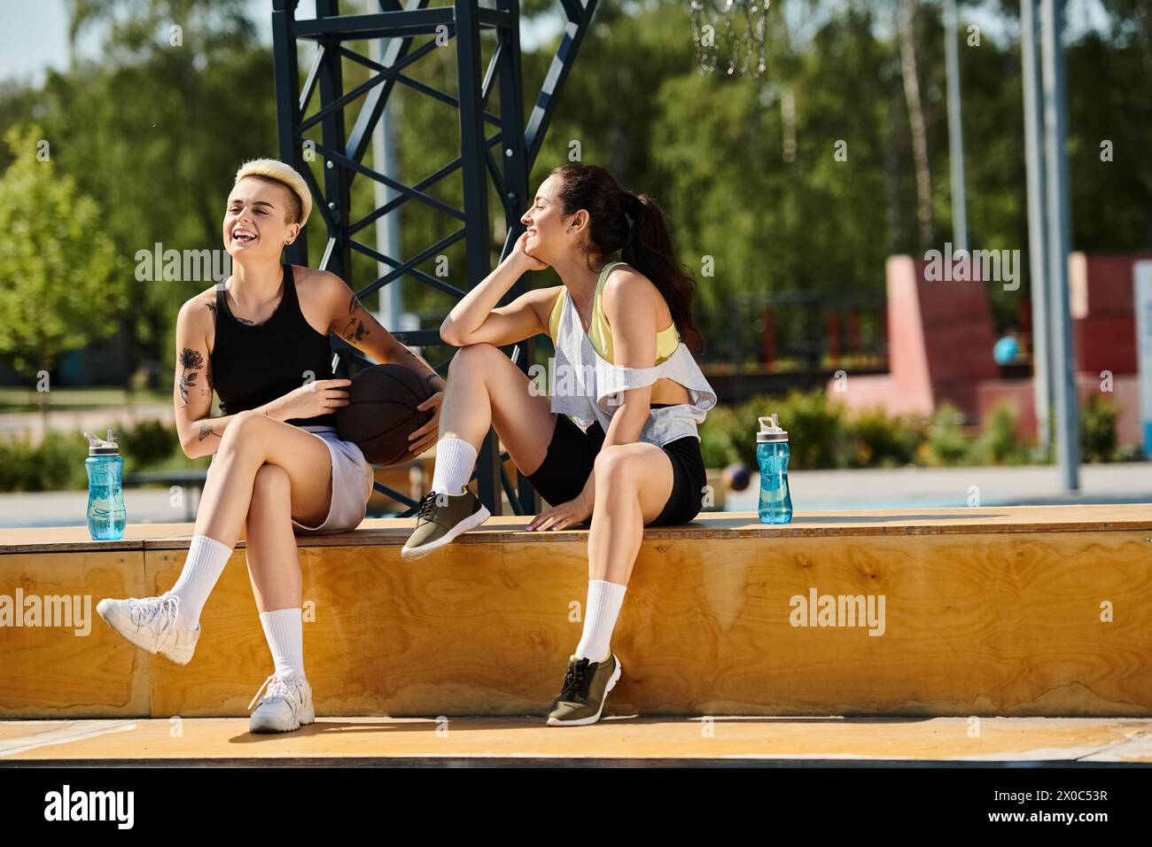 Two athletic young women sitting on a ledge, sharing a moment of joy and laughter while enjoying a summer day outdoors. Stock Photo