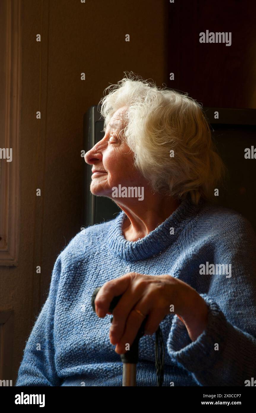 Portrait of old woman at home, smiling and looking at the window. Low key. Stock Photo