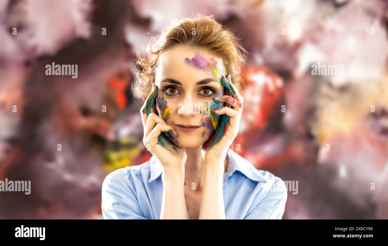 Woman Painter of abstract paintings painted with paints on her face, posing in front of her painting. Stock Photo