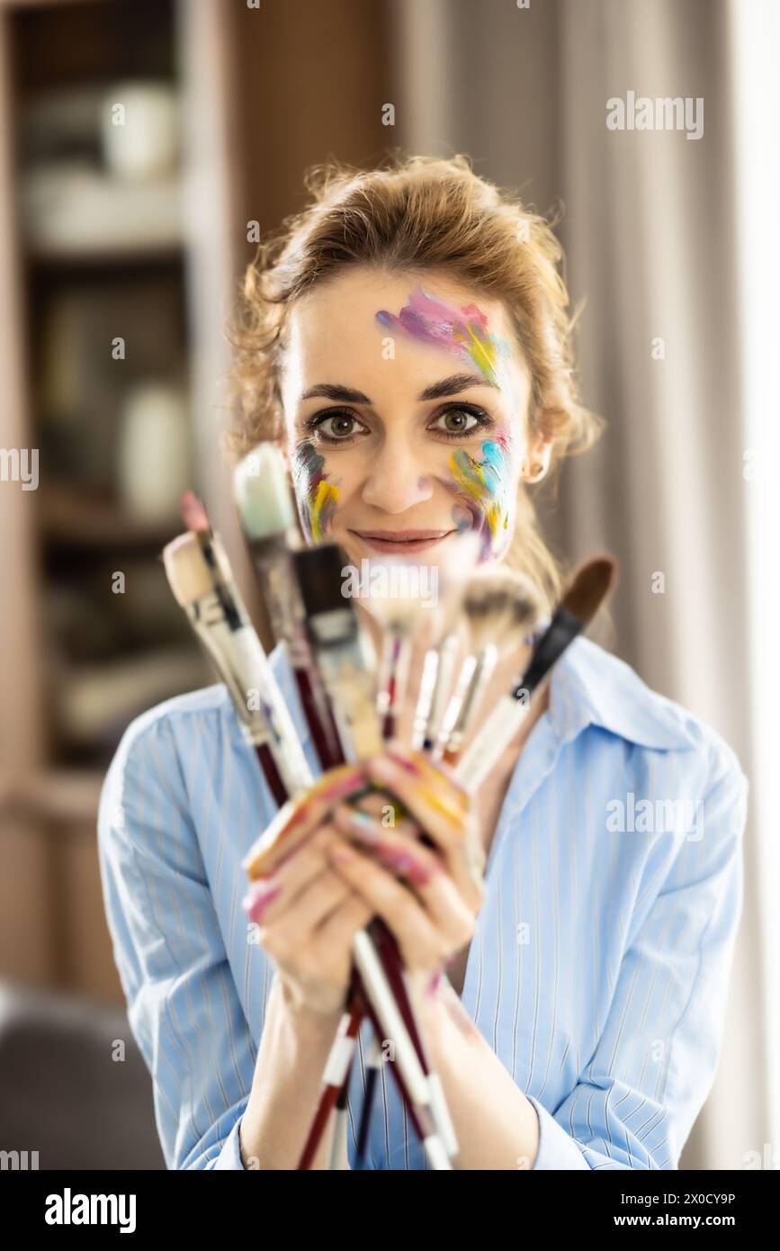 Portrait of a cute Latin woman holding a bunch of paintbrushes with both hands and smiling. Stock Photo