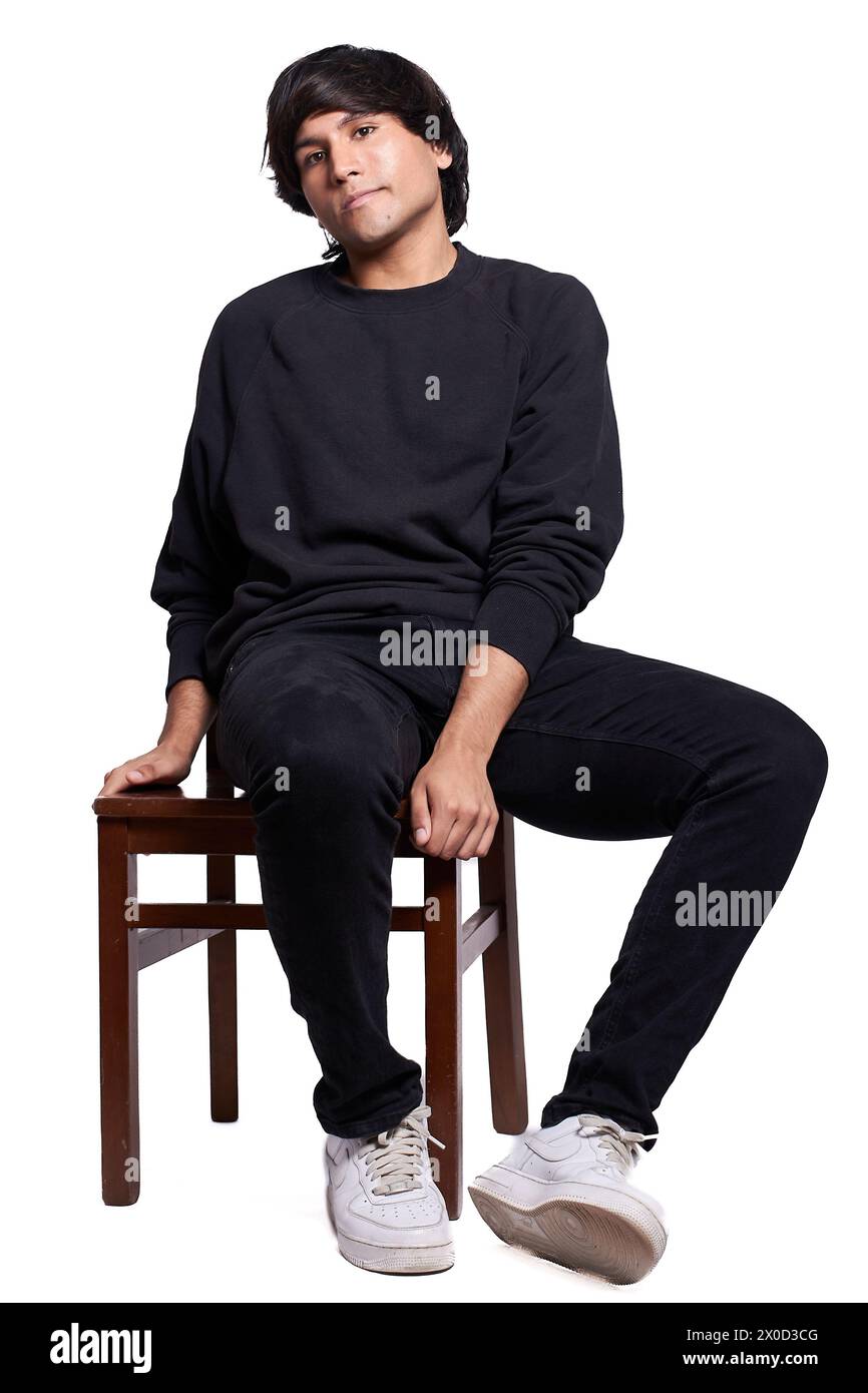 Latino man dressed in black is posing for a photo shoot. He is sitting on a wooden chair, the background is white. Stock Photo