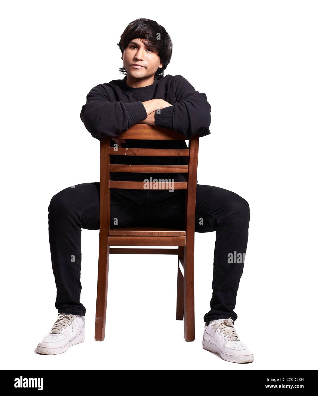 A man is sitting on a wooden chair with his arms crossed. He is wearing a black shirt and black pants. The chair is brown and has wooden slats. The ma Stock Photo