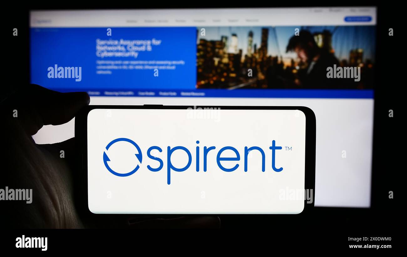 Person holding cellphone with logo of British company Spirent Communications plc in front of business webpage. Focus on phone display. Stock Photo