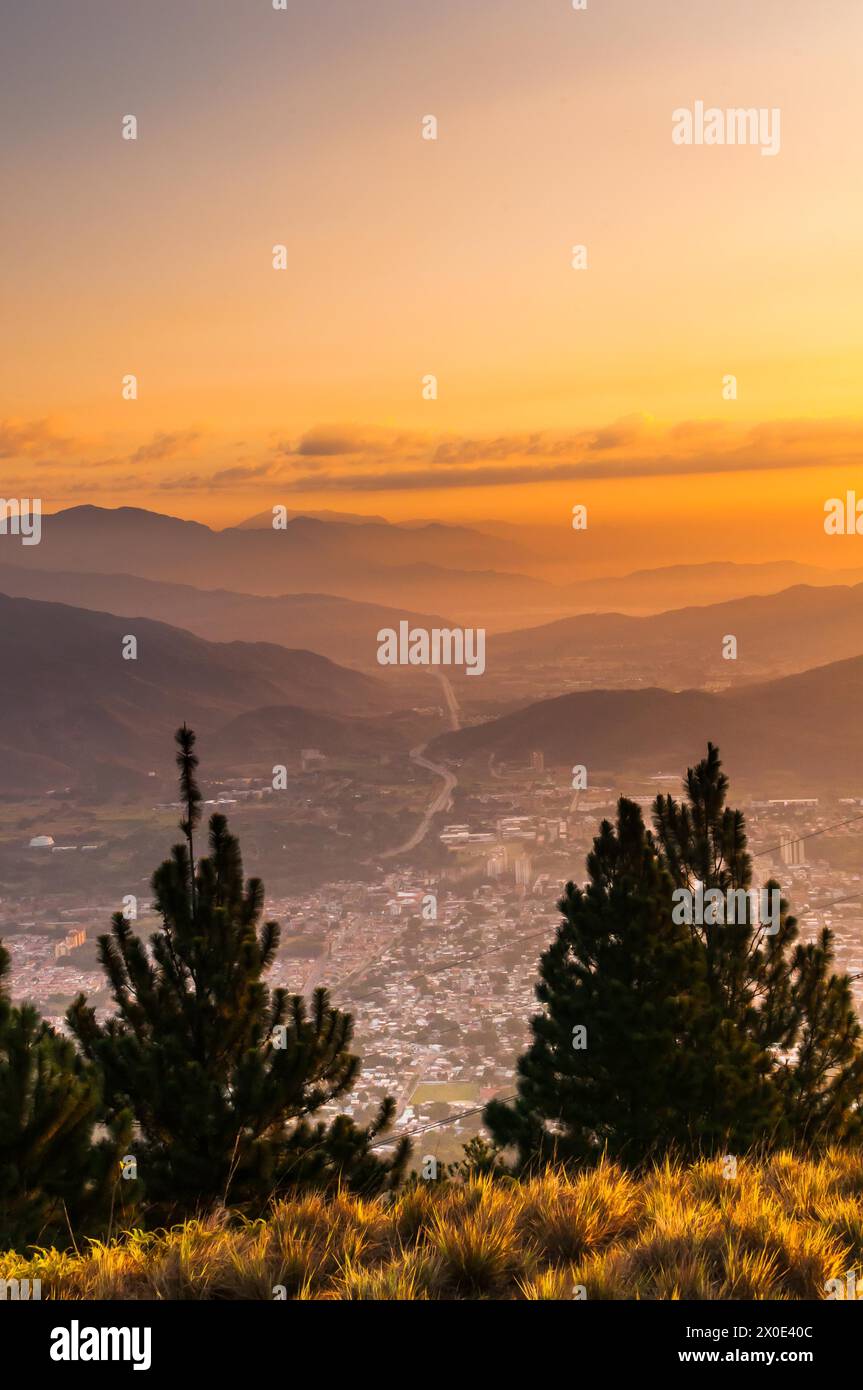 View from Cerro El Cafe of a sunrise from a mountain, where you can see a city and a highway that runs through it and several mountains. Stock Photo