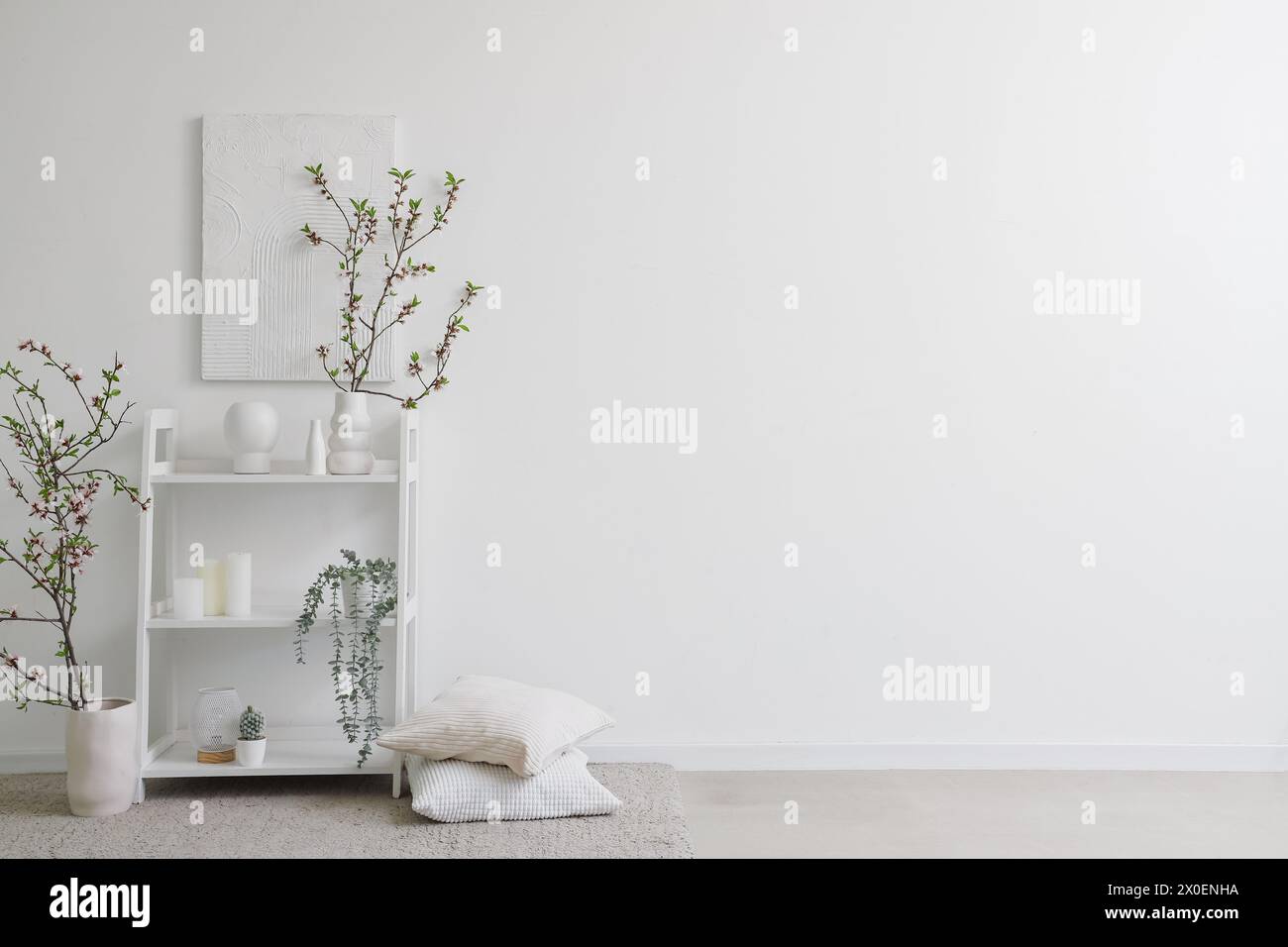 Vases with blooming branches and decor on shelving unit near white wall Stock Photo