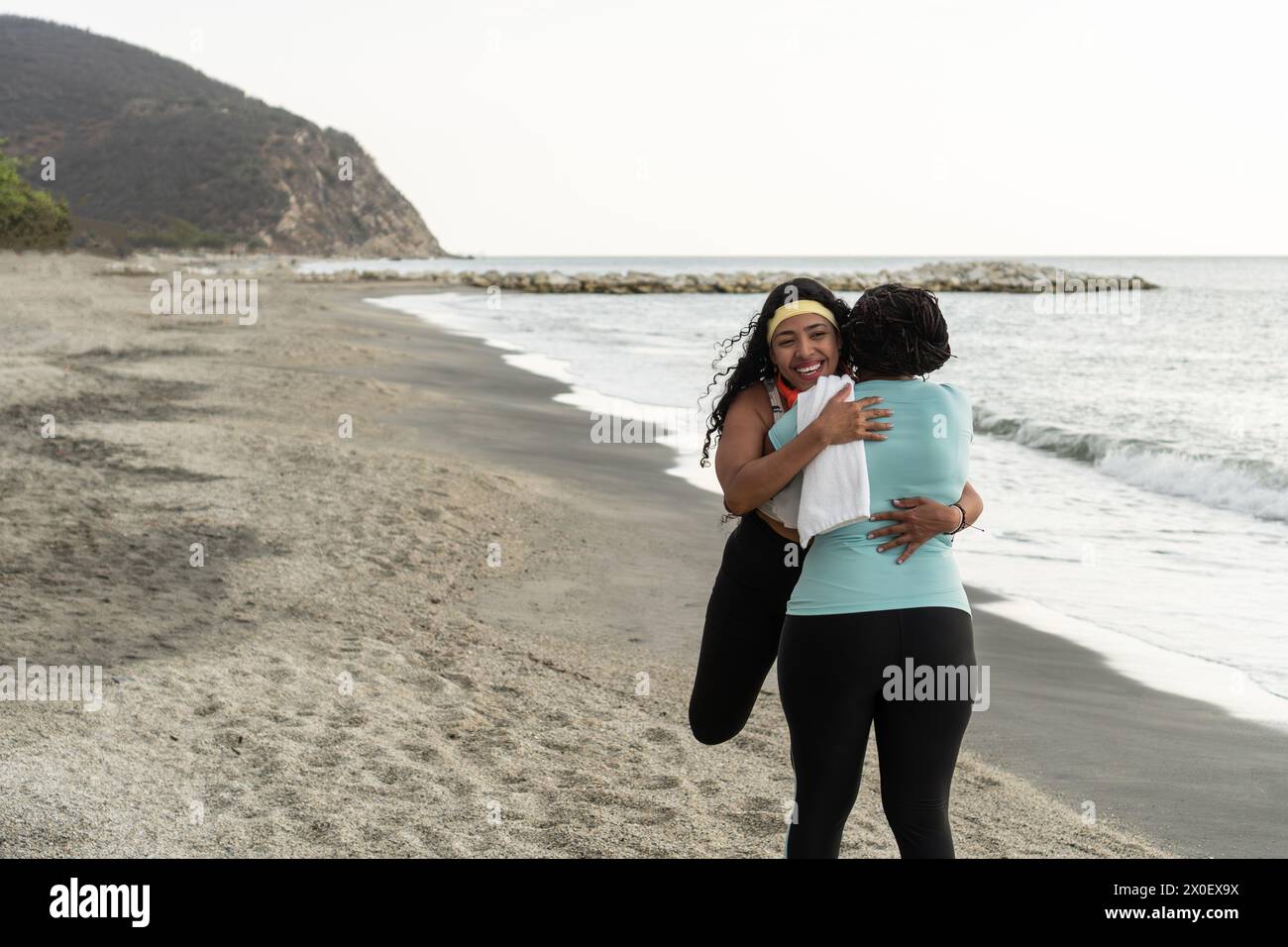 Two women embrace joyfully on a quiet beach, waves gently lapping the shore. Stock Photo