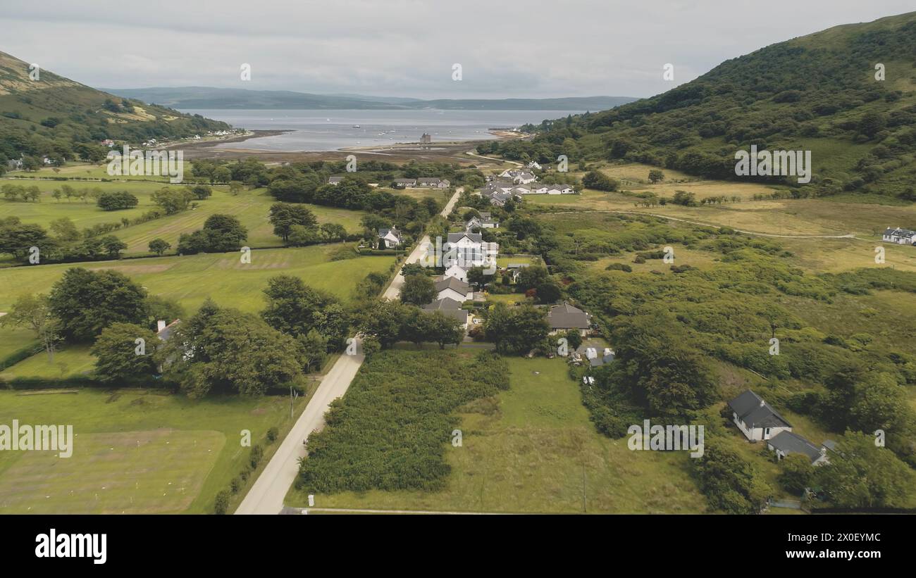 Slow motion of Scottish whisky distillery at village aerial. Road, cottages, houses at green mountain valley. Summer cloudy day. Nobody nature landscape of Arran Island, Scotland. Cinematic drone shot Stock Photo