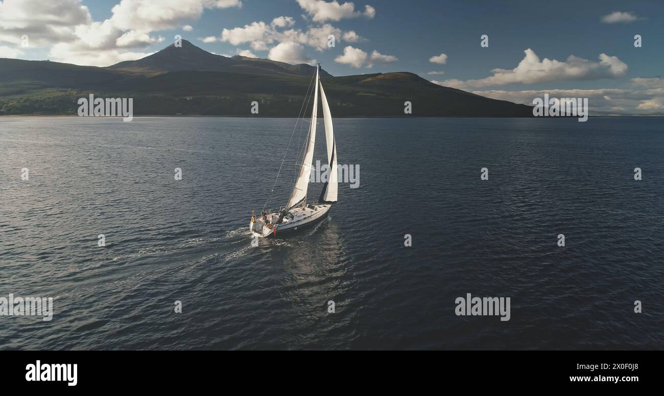 Slow motion yacht sails at mountain island shore aerial. White sailboat at Arran isle, Scotland, Europe. Sun shine above mounts with dark forest at cloudy summer day. Cinematic cruise on boat Stock Photo