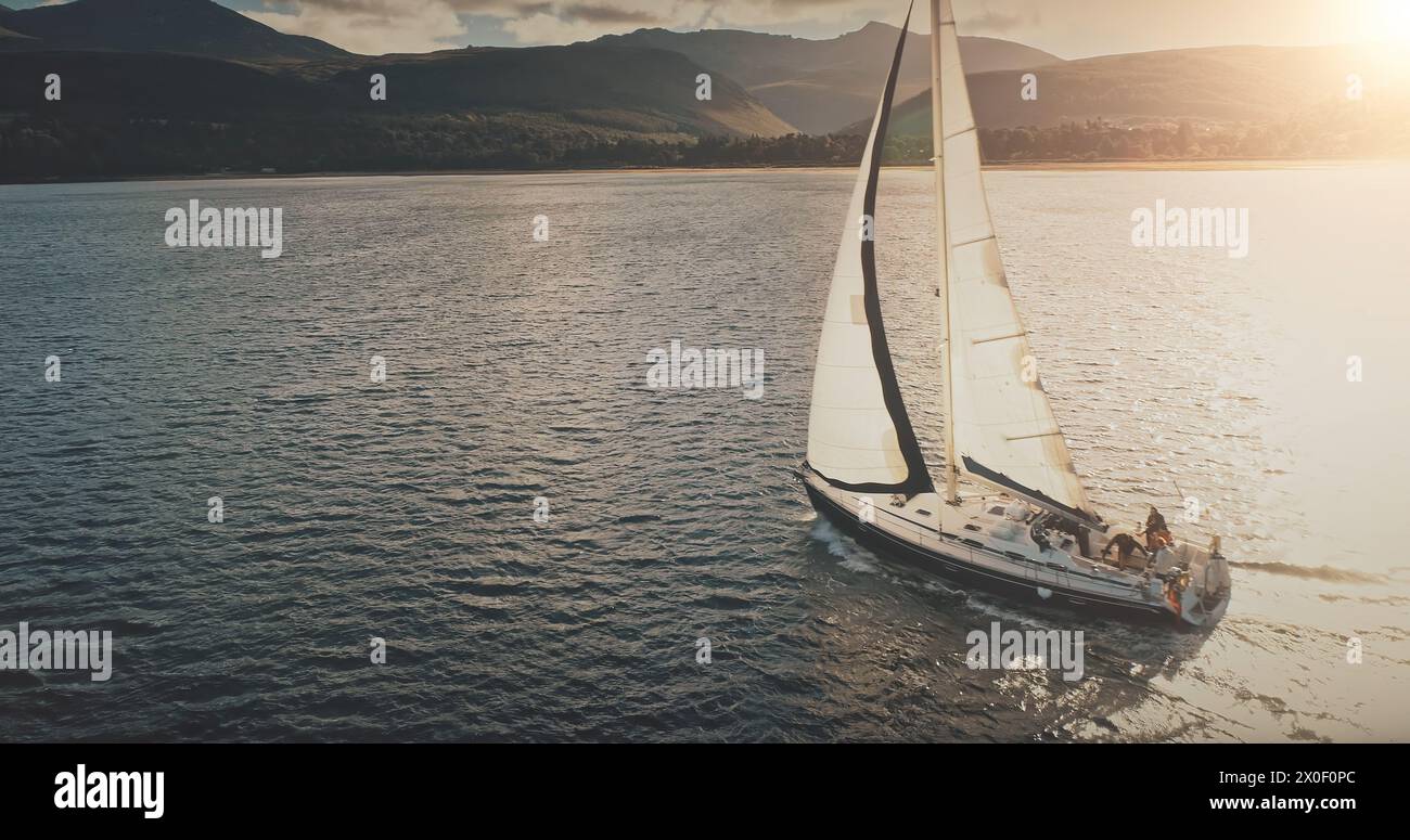 Sun shine over yacht at mountain island coast aerial. Luxury sailboat regatta at sea gulf. Sunlight flare reflection at serene seascape. Tourist cruise at Brodick harbor, Arran islet, Scotland, Europe Stock Photo