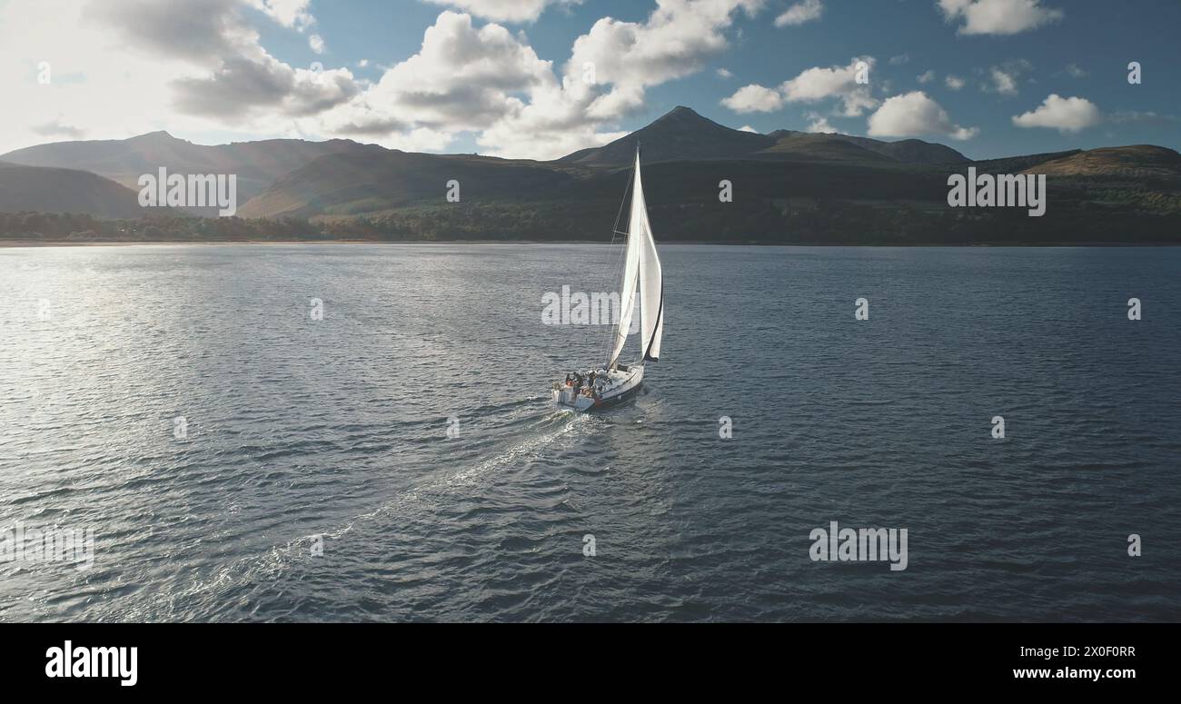 Slow motion of yacht, wind blows to sail at ocean bay aerial. Mountain island of Arran, Scotland, Europe. Nobody nature seascape at Brodick Gulf. Luxury cruise on sailboat. Cinematic tourism lifestyle Stock Photo