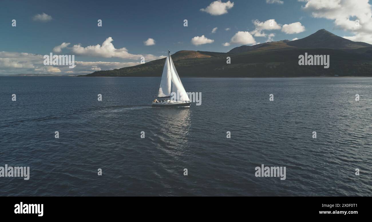 Slow motion of yacht, wind blows to sail at ocean bay aerial. Mountain island of Arran, Scotland, Europe. Nobody nature seascape at Brodick Gulf. Luxury cruise on sailboat. Cinematic tourism lifestyle Stock Photo