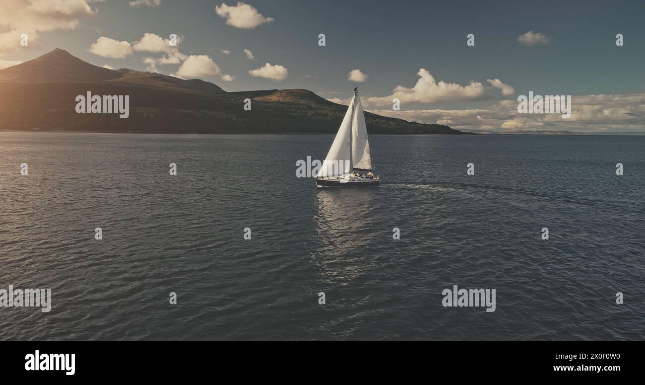 Sail boat at mountain ocean shore of Scotland Island of Arran aerial. Yacht sails with beautiful fluffy clouds sky at open sea. Sailboat cruise at summer sun shine day. Cinematic soft light drone shot Stock Photo
