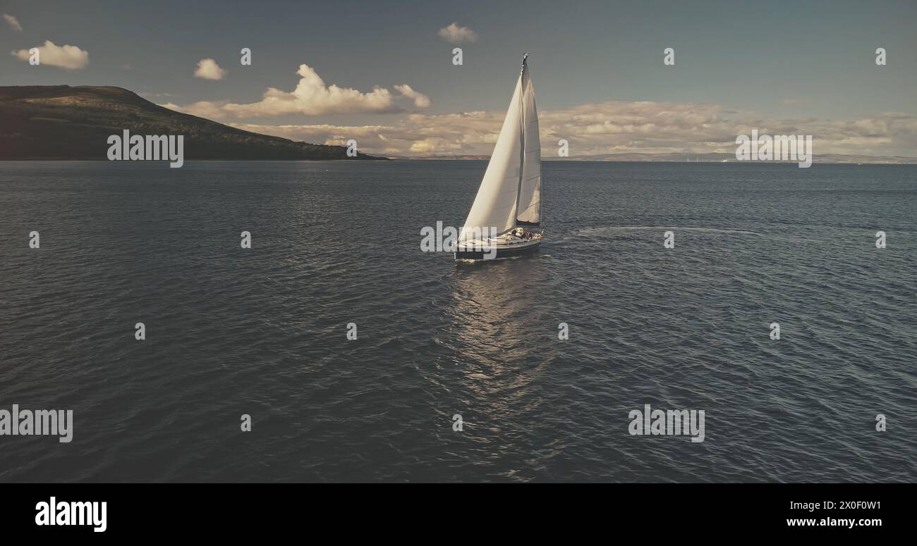 Sail boat at mountain ocean shore of Scotland Island of Arran aerial. Yacht sails with beautiful fluffy clouds sky at open sea. Sailboat cruise at summer sun shine day. Cinematic soft light drone shot Stock Photo