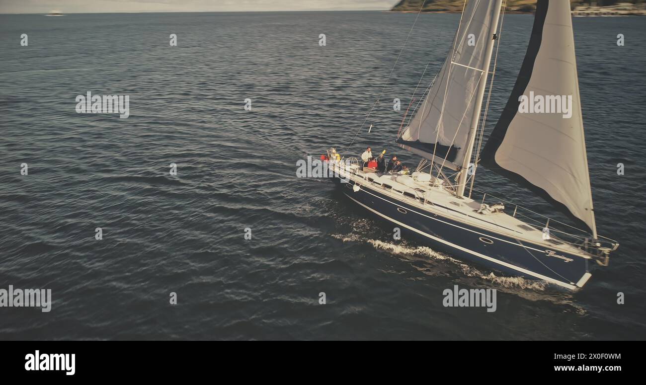 Sail boat at mountain ocean shore of Scotland Island of Arran aerial. Yacht sails with beautiful fluffy clouds sky at open sea. Sailboat cruise at summer sun shine day. Cinematic soft light drone shot Stock Photo