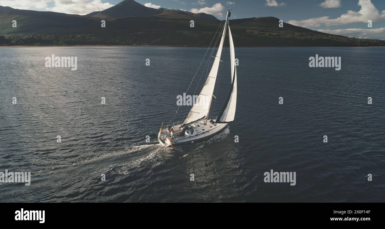 Yacht cruise in sunlight reflection at open sea bay aerial. Luxury sailboat sailing at Brodick Pier, Arran Island, Scotland. Mountain isle on ocean shore. Cinematic serene seascape with lonely ship Stock Photo
