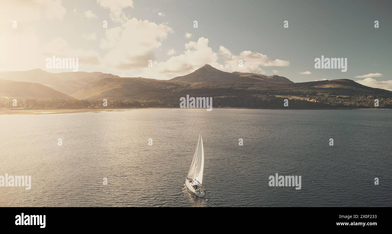 Sun reflection at ocean bay with sailboat. Aerial yacht cruise at sunlight. Summer luxury journey on sail boat at open sea. Mountain island of Arran, Scotland at cinematic seascape at soft drone shot Stock Photo