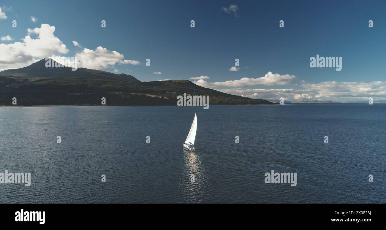 Aerial racing yacht at mountainous island. Luxury ship at mountain ocean coast. Sailboat regatta at sea bay. Sun reflect at Brodick pier, Arran isle, Scotland, Europe. Sail boat summer cruise Stock Photo