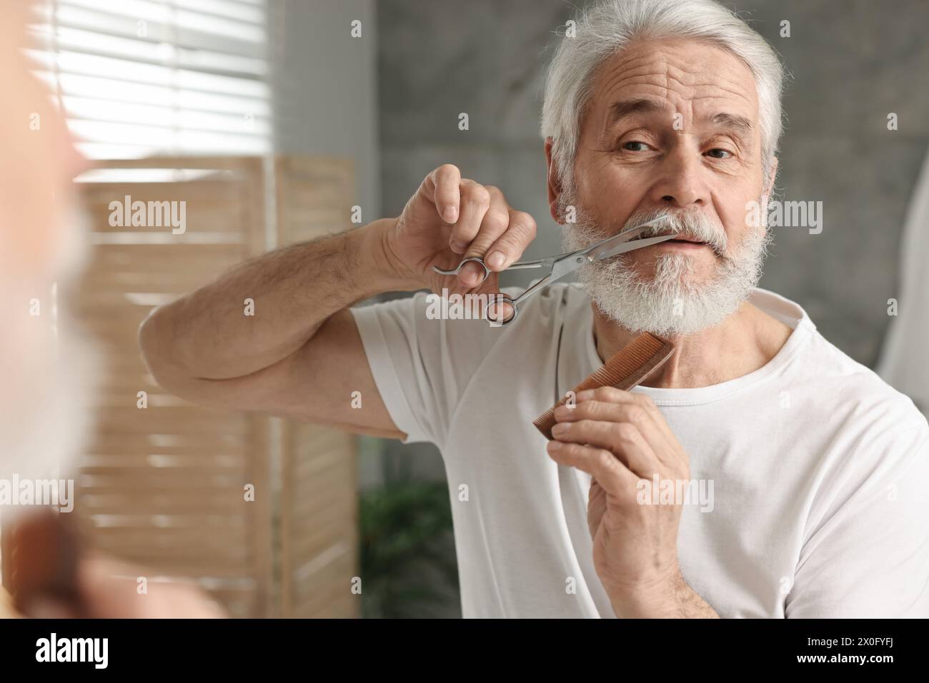 Senior man trimming mustache with scissors near mirror in bathroom