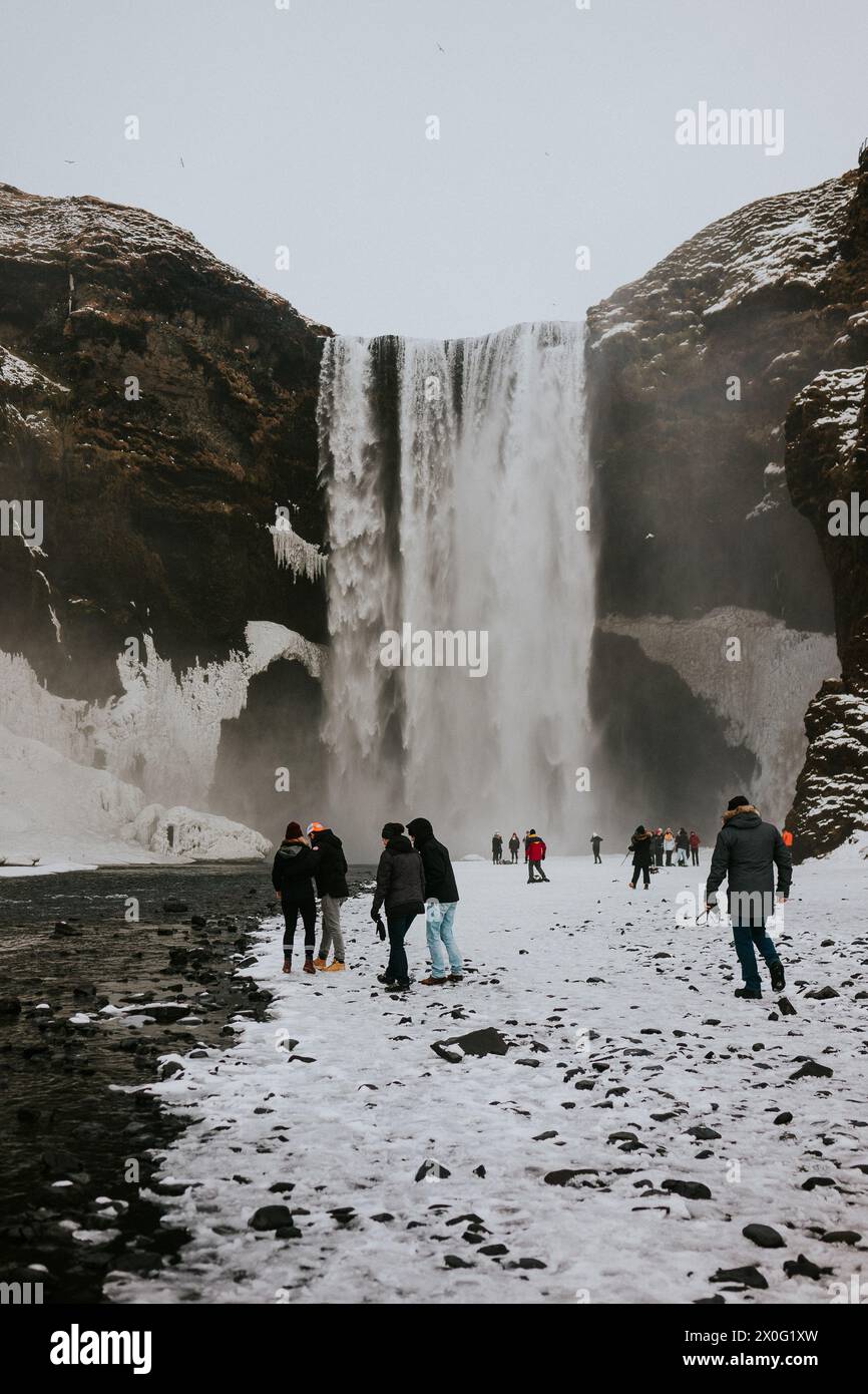 Tourists in outerwear walk by the Skoga river and Skogafoss waterfall Stock Photo
