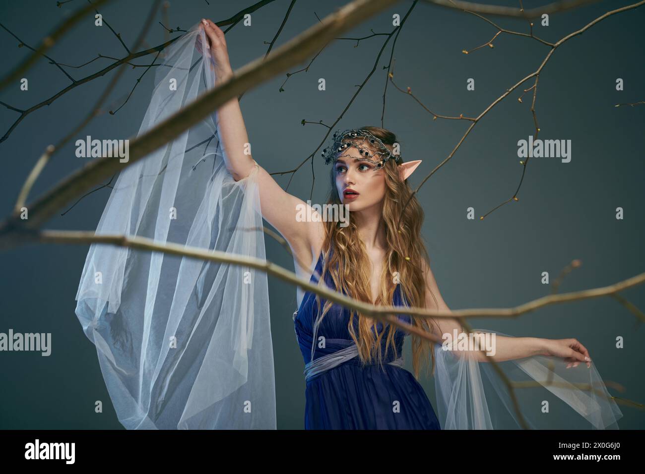 A young woman in a blue dress gracefully holds a delicate white veil in a magical studio setting fit for an elf princess. Stock Photo