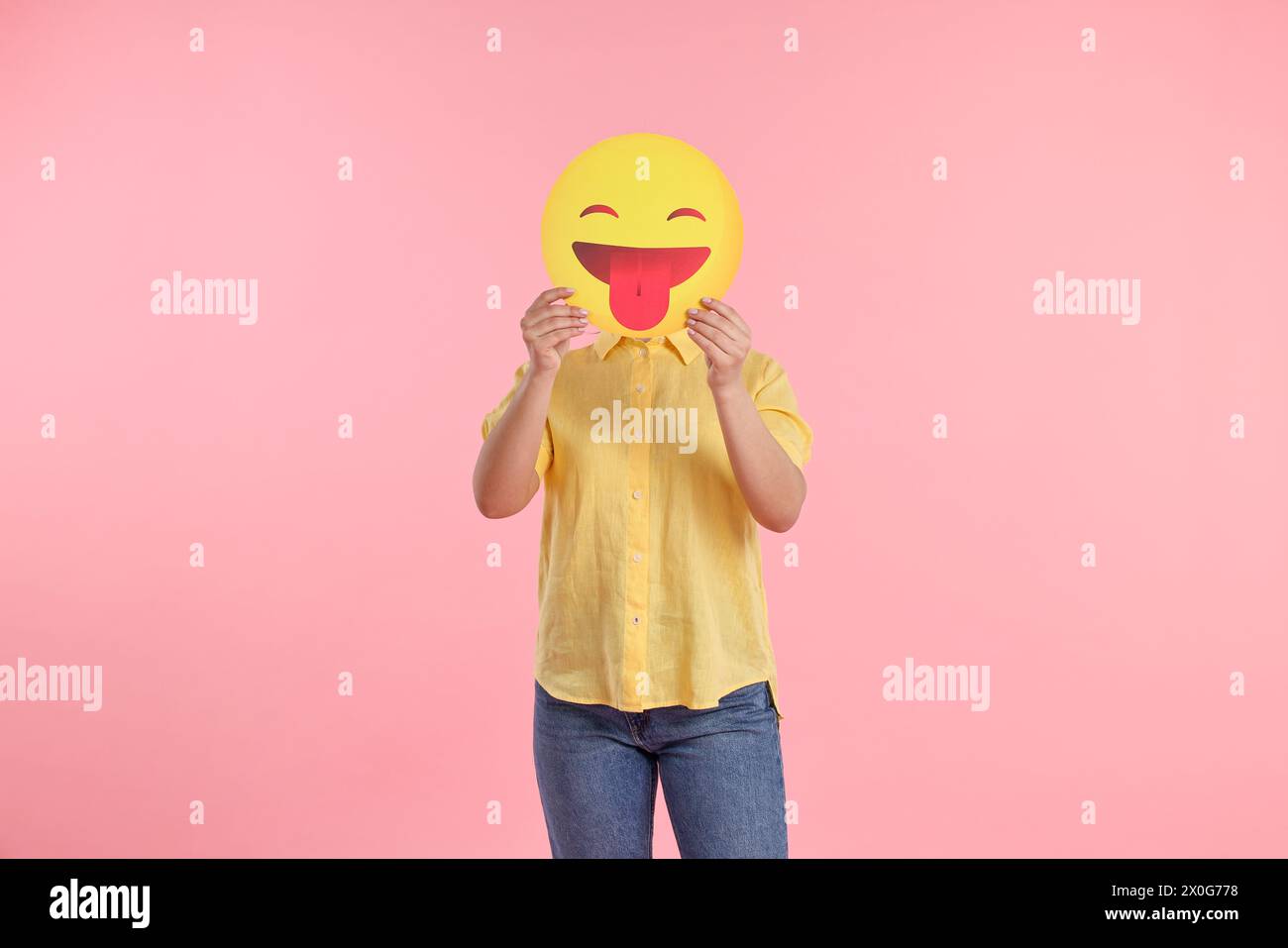 Woman covering face with emoticon sticking out tongue on pink background Stock Photo