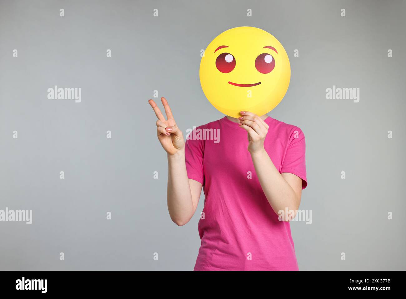 Woman covering face with smiling emoticon and showing peace sign on grey background. Space for text Stock Photo