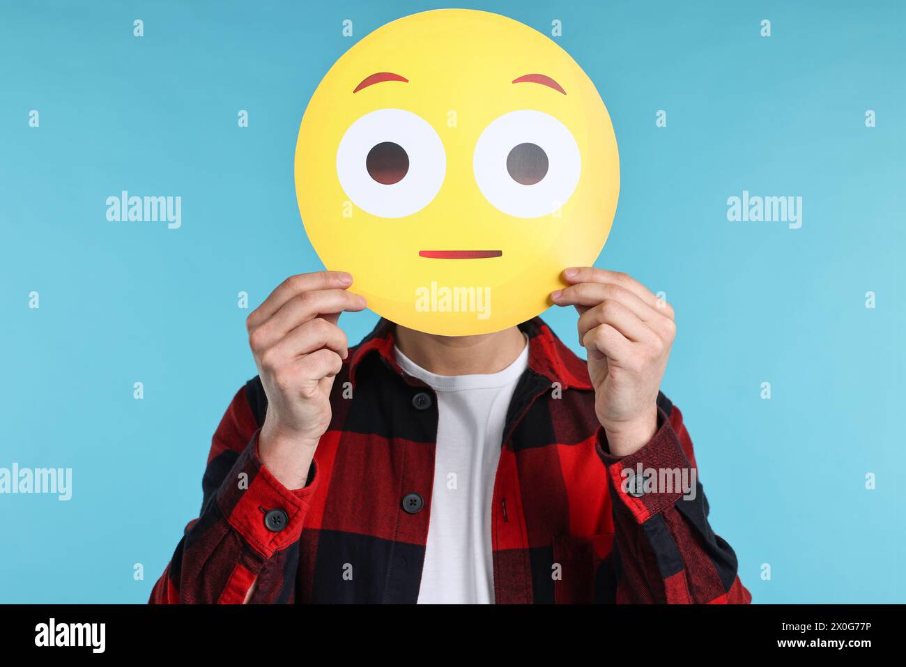 Man covering face with surprised emoticon on light blue background Stock Photo