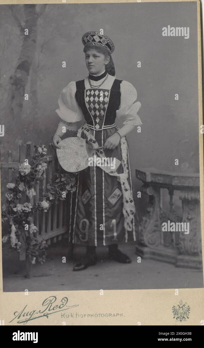 'Photograph of Mizzi Beutel (approx. 20 years old) in carnival costume Studio shot with fence decorated with flowers and terrace balustrade Clothing consisting of hood with playing cards, white blouse, long dress with rhombuses at the bust, apron with playing cards and numbers ''9'' and ''1'', loose belt with dominoes, carrying a fan in her hand [automated translation]' Stock Photo