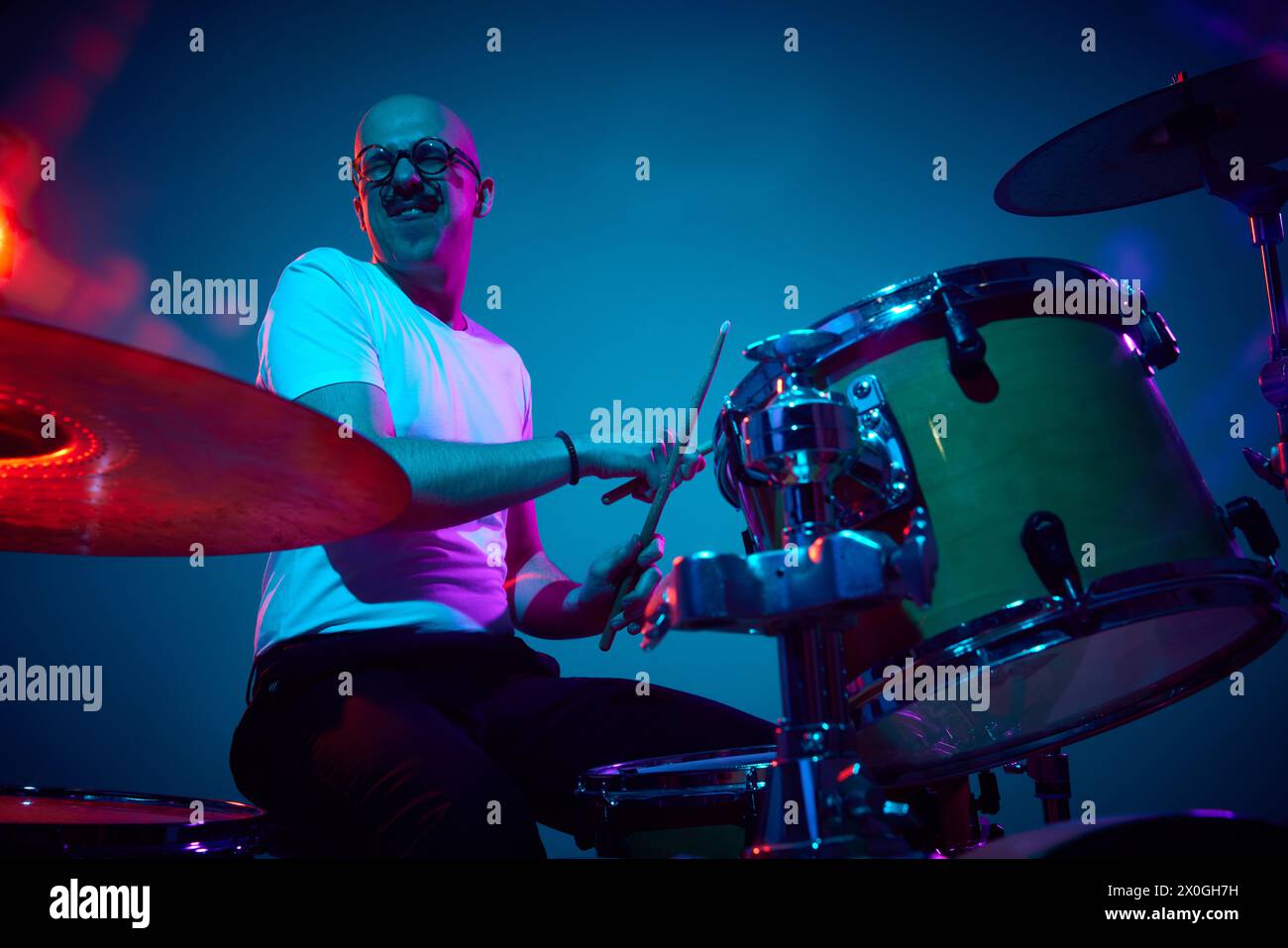 Artistic bald drummer in casual outfit playing in pink-purple stage ...