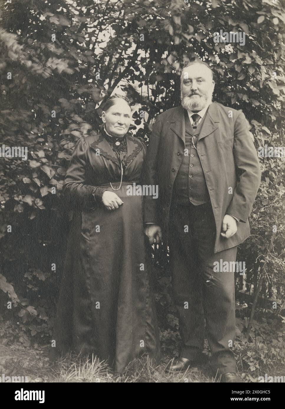 A couple of about 70 years old are standing close together. He is holding her left hand with his. They both have a good belly, on different parts of their upper bodies. They are both wearing black clothes, she a floor-length dress with a lace trim and a necklace with a medallion, her hair braided into a wreath, he a three-piece suit, the jacket open, the vest stretched around her stomach. He is a head taller than his rouges and has a full, well-trimmed beard. They are standing in a garden in front of a tall bush [automated translation] Stock Photo