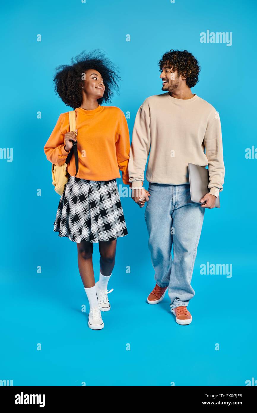 An interracial couple, casually dressed, takes a leisurely walk together against a vibrant blue backdrop in a studio setting. Stock Photo