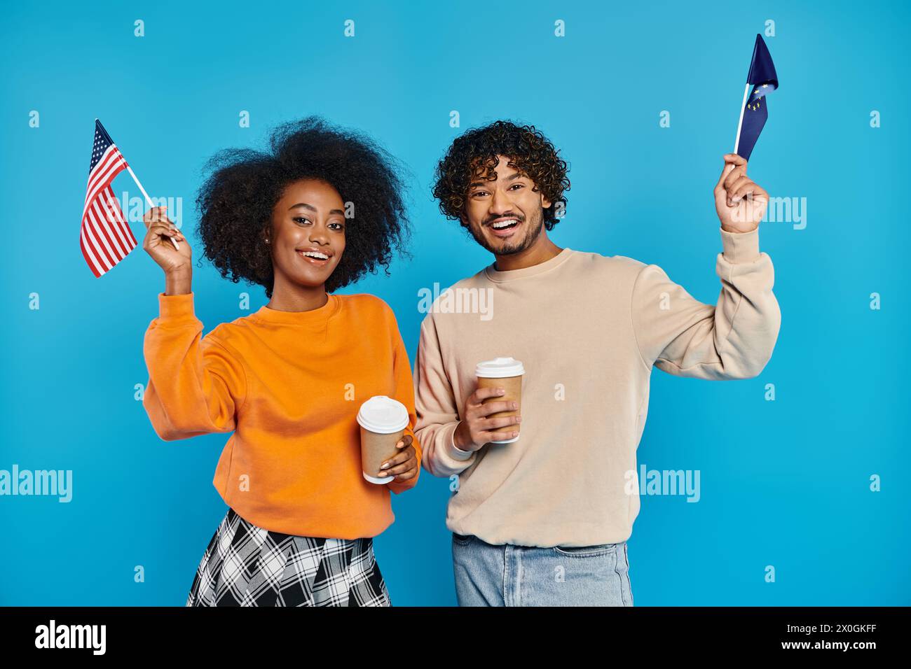 An interracial couple, dressed casually, proudly hold American flags against a blue backdrop. Stock Photo