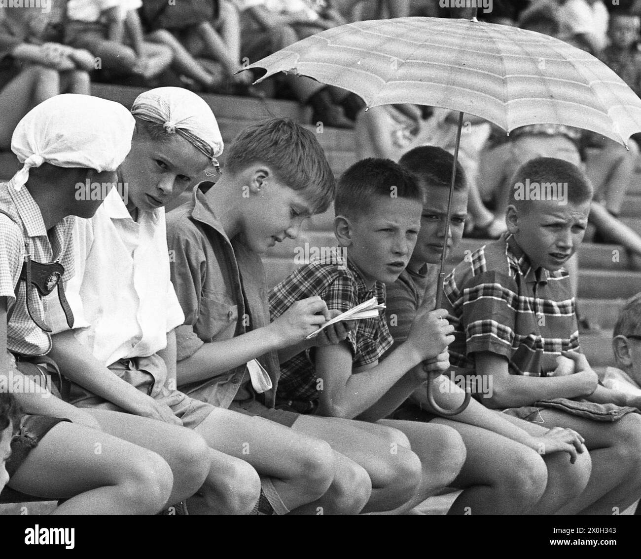 Sports-loving children watch a game in a stadium. A boy holds an umbrella to protect himself from the sun. [automated translation] Stock Photo