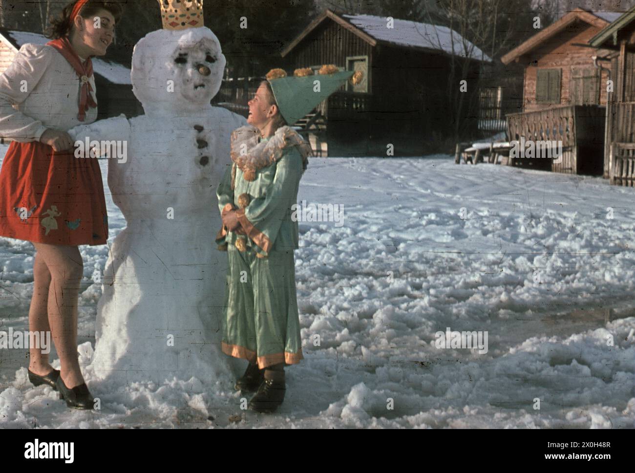 Two girls in carnival costumes have built a snowman with a crown at the Wörthsee. The girl on the right is wearing a clown costume. In the background there are houses. [automated translation] Stock Photo