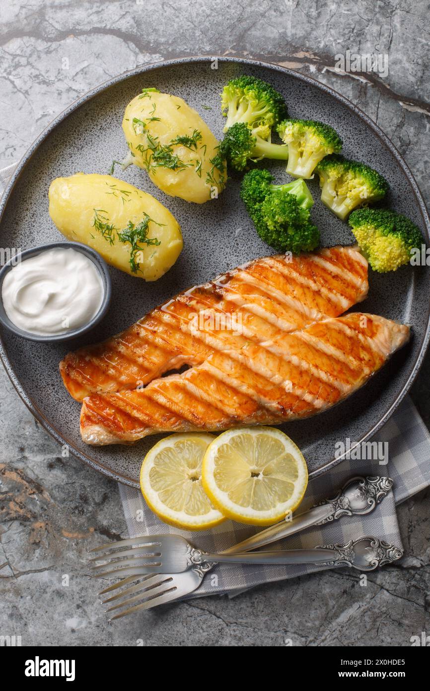 Grilled salmon steak with new potatoes, broccoli and cream sauce close-up in a plate on the table. Vertical top view from above Stock Photo