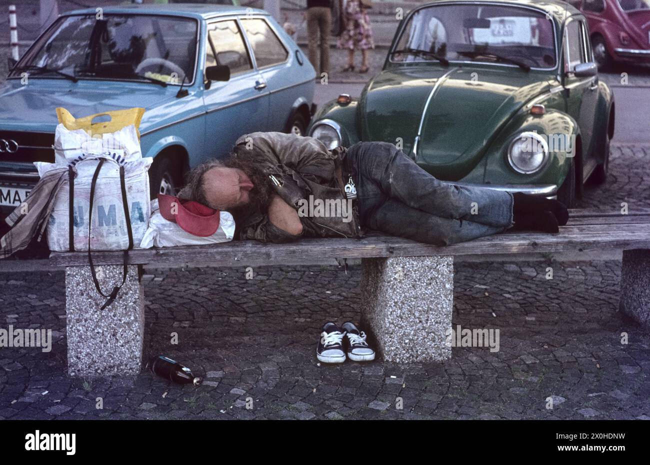 A homeless man sleeps on a bench in front of the Munich University in Schwabing. he has put his shoes under the bench. Next to him his things are in simple bags, on the floor is a bottle. Behind him are some cars, a turquoise Audi and a dark green VW Beetle. [automated translation] Stock Photo