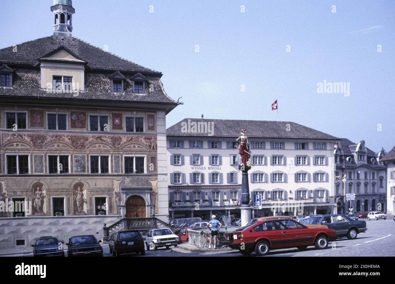 Cars are parked on the main square. A cyclist with a racing bike stands at the fountain with the Brunnen-Mändl. The town hall is on the left and the Hotel Wysses Rössli is at the back right [automated translation] Stock Photo