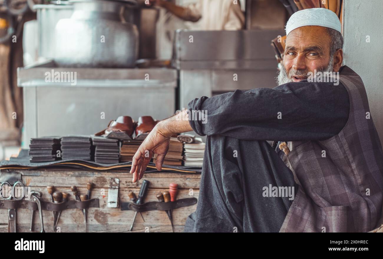 Poor old sad Pakistani Pathan shoe man cobbler on the local streets of Pakistan with his hand made leather shoes and repair tools in his street shop Stock Photo