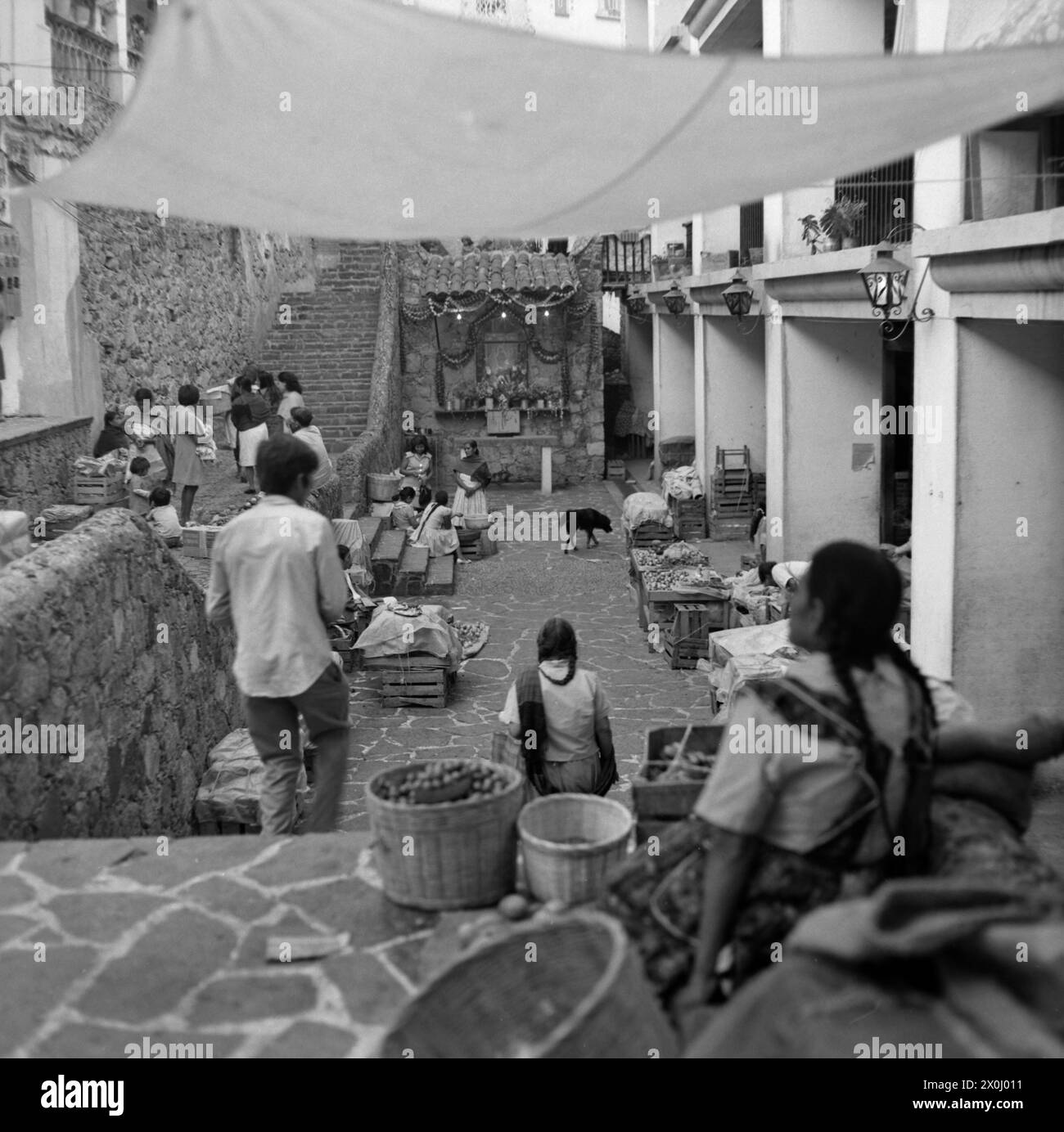 View into an inner courtyard in Taxco. There are many young people in the courtyard. Some are sitting on stone stairs. There are many wooden boxes standing around. At the other end of the courtyard there is a small shrine. Above the courtyard there are cloths stretched between the buildings. [automated translation] Stock Photo