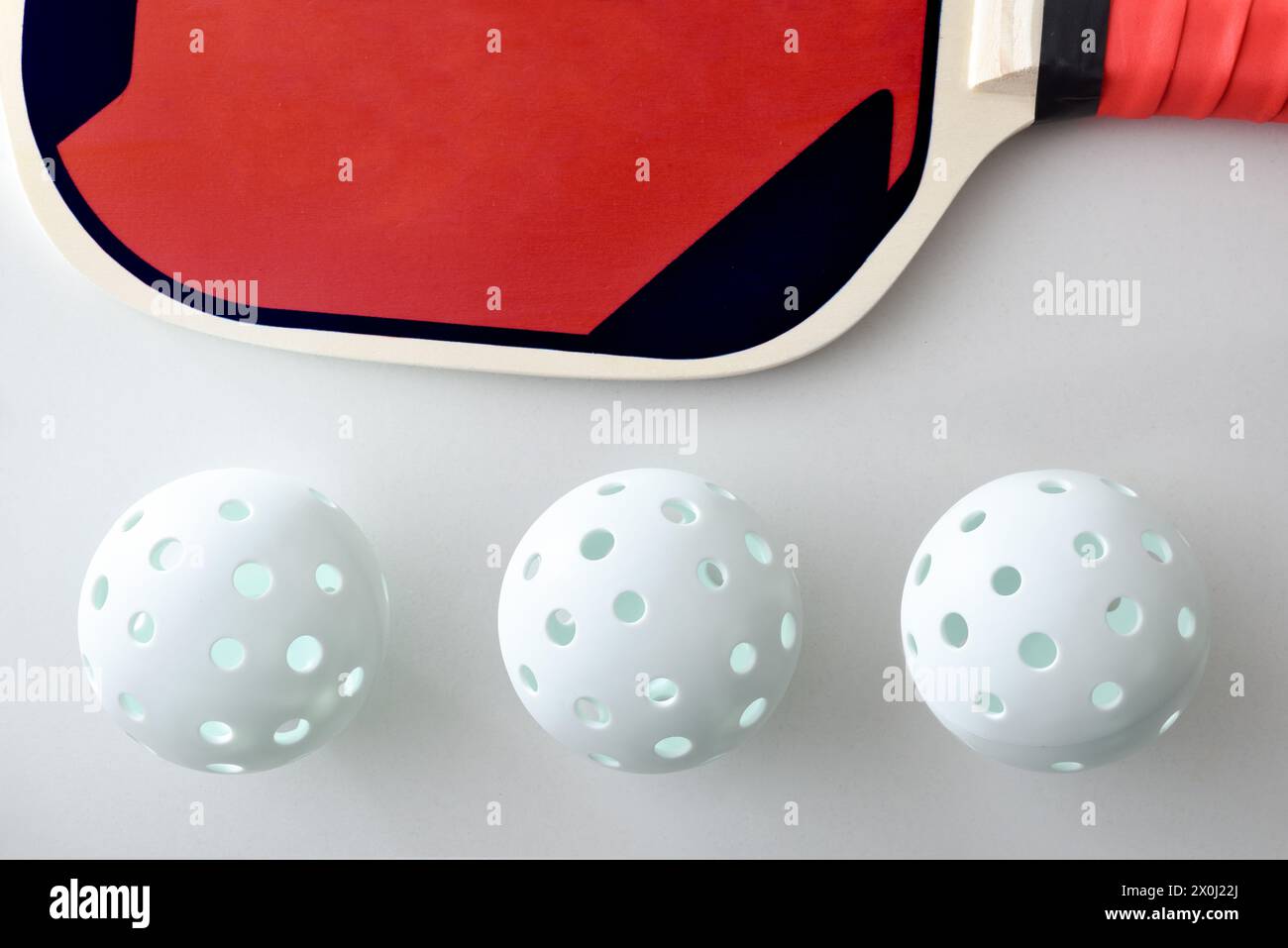 Detail of three lined up pickleball balls and red and black wooden paddle isolated on white table. Top view. Stock Photo