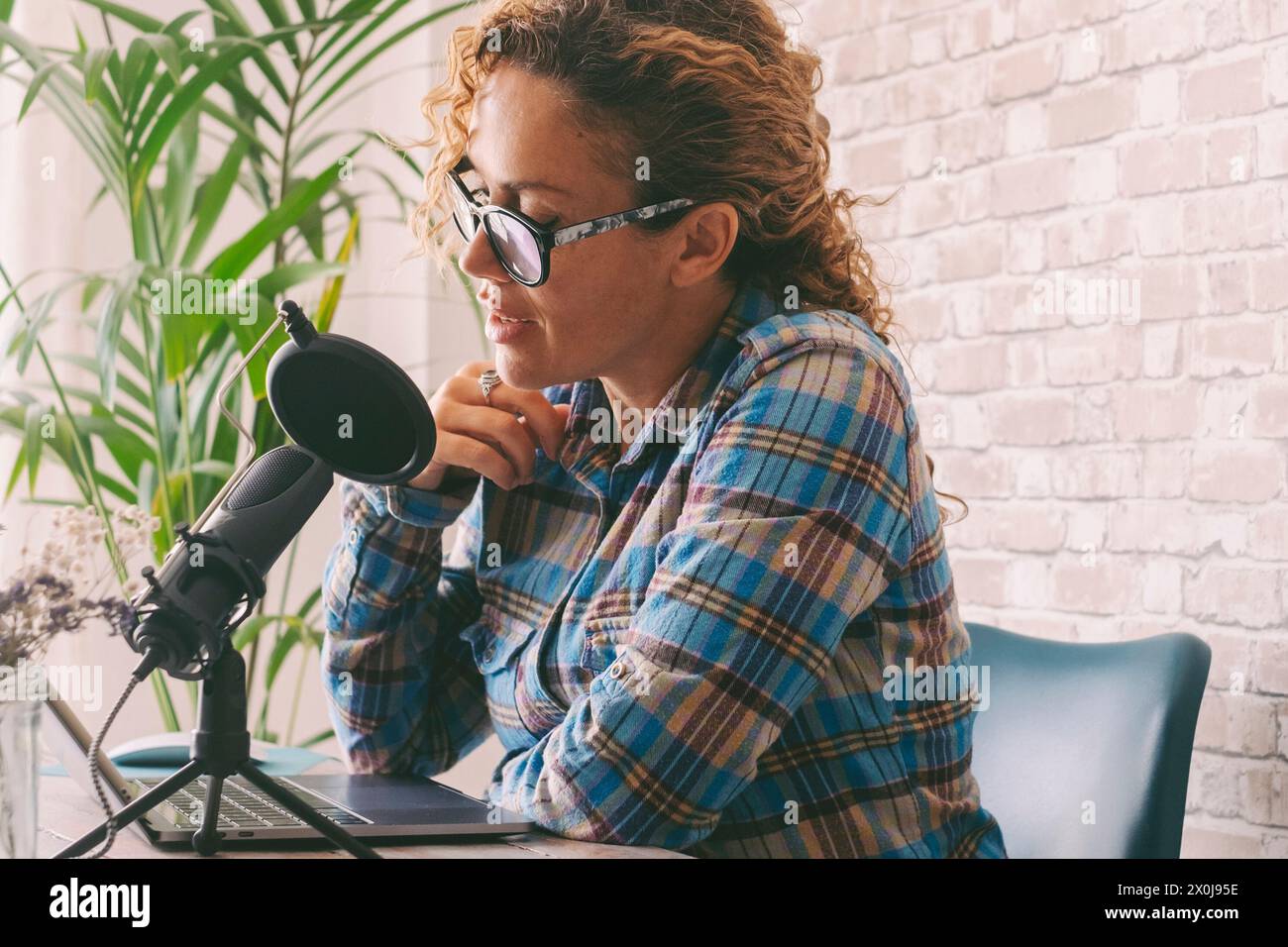 Content creator adult woman recording a podcast interview using microphone and laptop sitting at the desk. Home office workstation and smart working modern people lifestyle. Online radio computer job Stock Photo