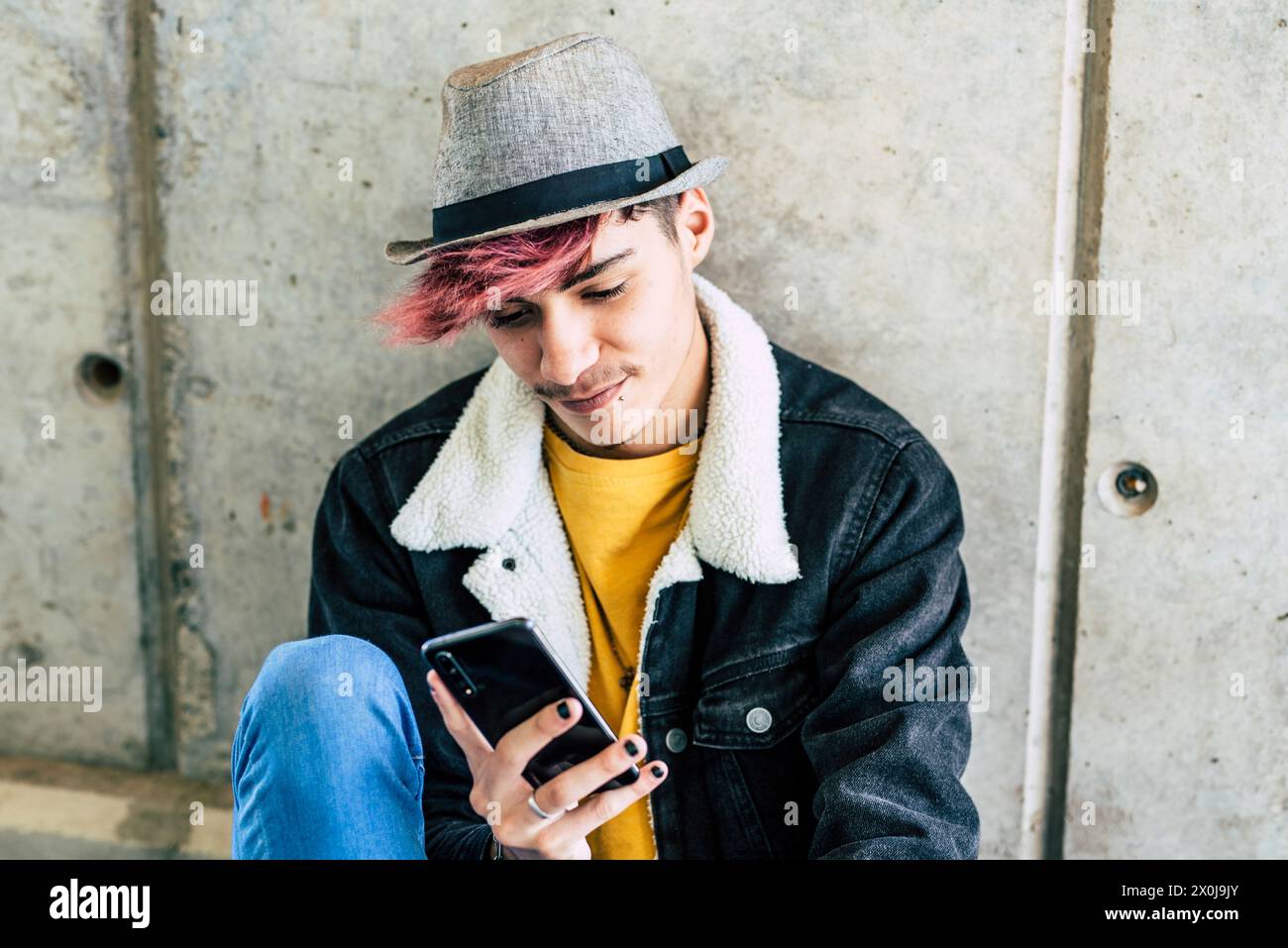 One young modern teenager sitting on the ground using phone app to message and social media life. Online connected youth male boy with trendy fashion alternative clothes in urban life background Stock Photo