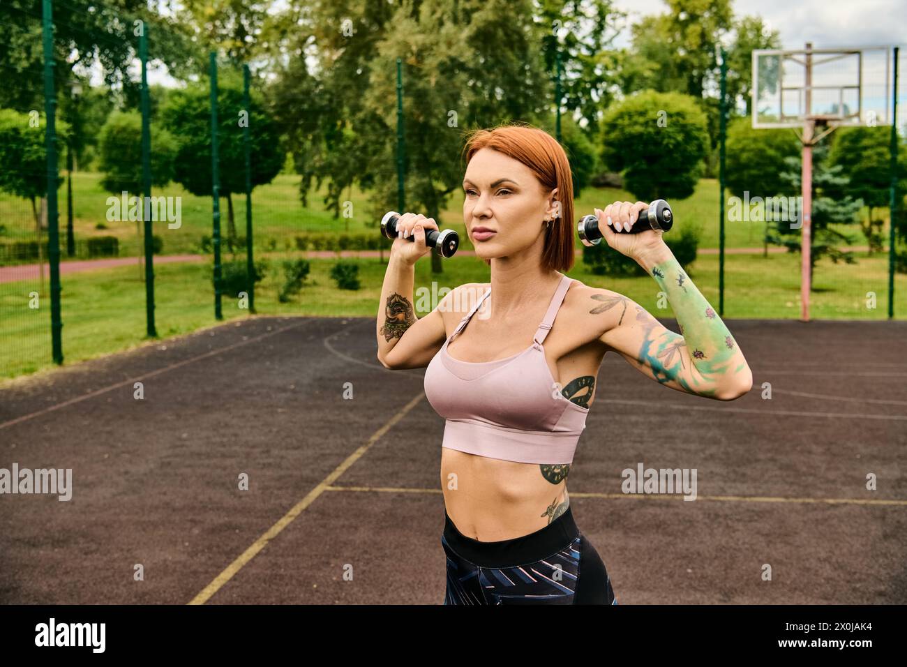 A woman in sportswear, holds a pair of dumbbells with determination and focus during outdoor exercise. Stock Photo