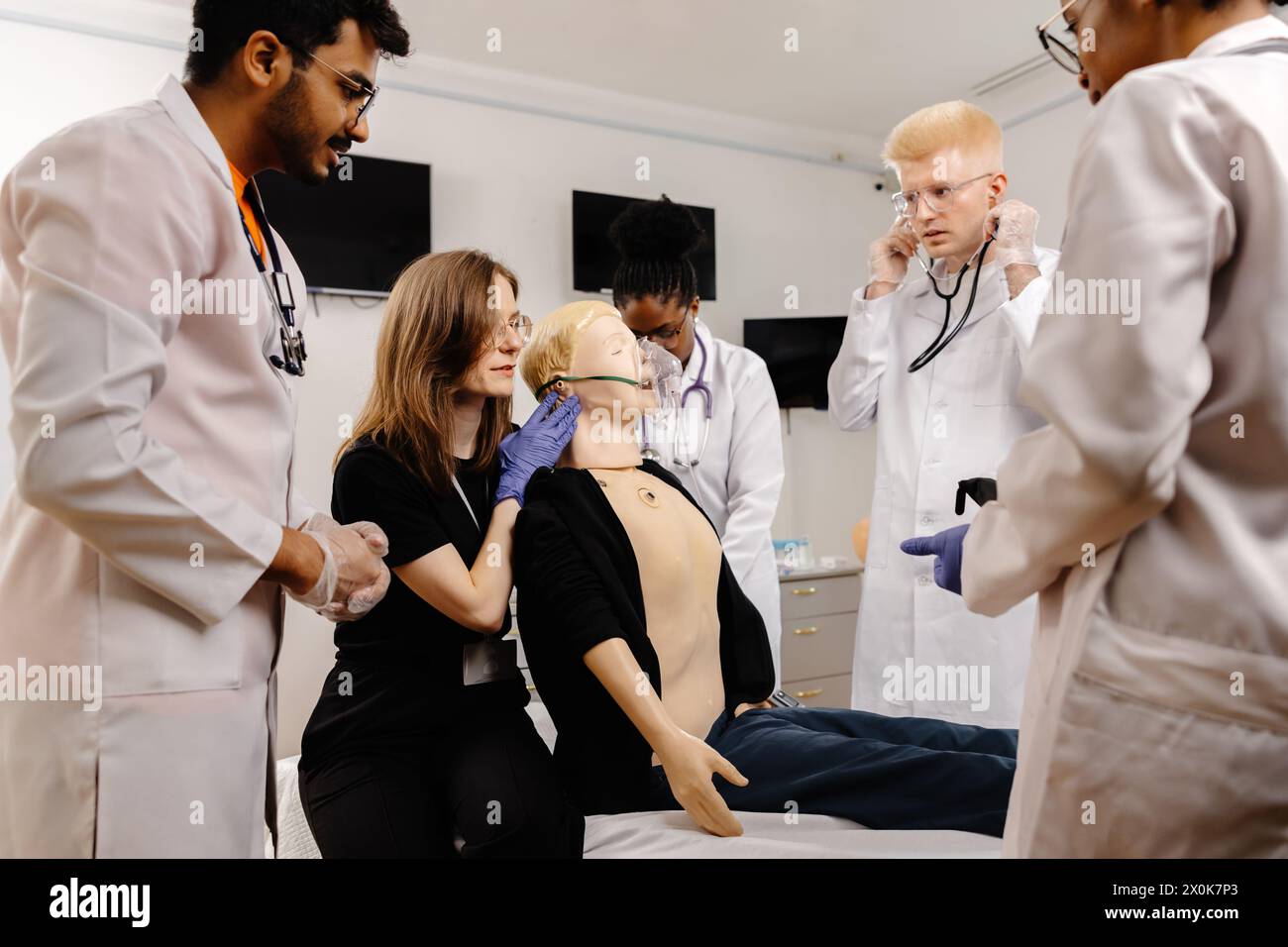 A group of doctors, wearing lab coats and stethoscopes, are gathered around a medical dummy, discussing and examining it in a hospital setting. Stock Photo