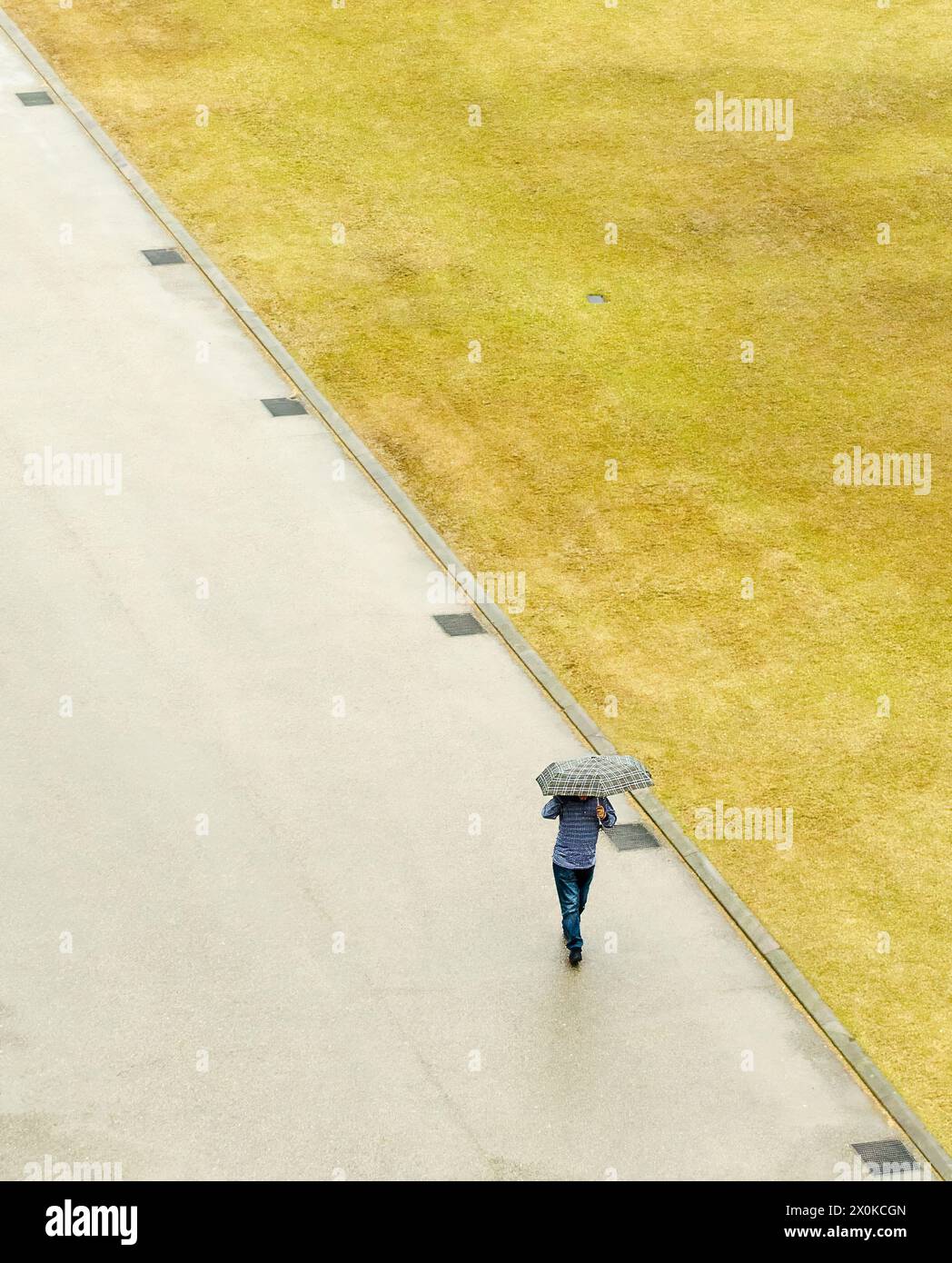 A person is walking down a sidewalk with an umbrella. The sky is cloudy and the grass is dry Stock Photo